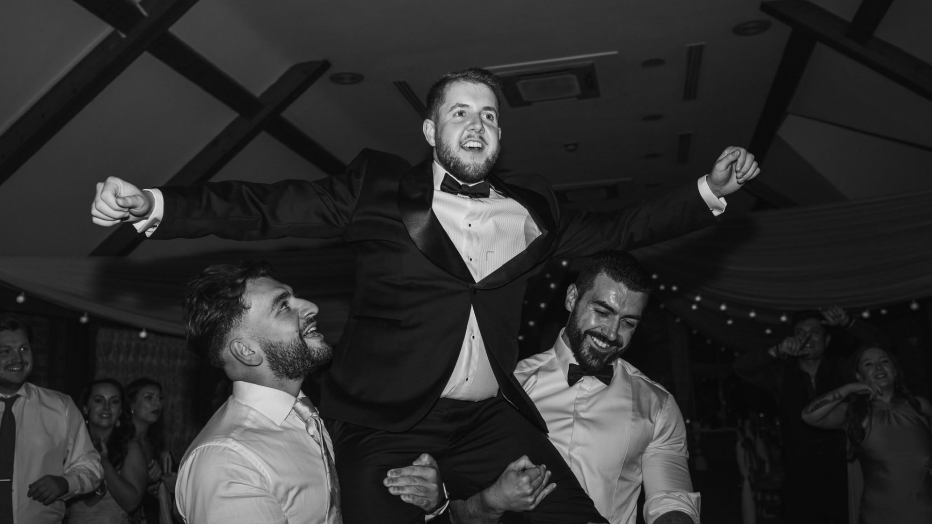 Black and white photo of Jared in his bespoke shawl-collar tuxedo by RN Tailoring, lifted onto the shoulders of his friends on the dance floor at his wedding